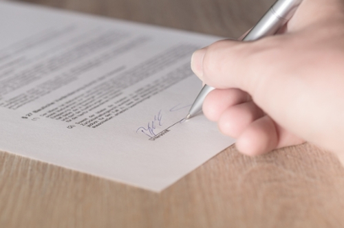 A person signs a piece of paper on a dotted line located at the bottom of the page.